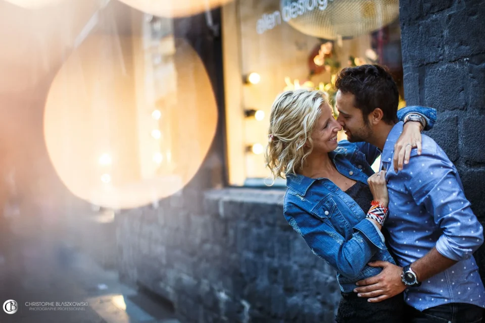 Couple enlacé dans le Vieux Lille, avec un effet de bokeh chaud en premier plan et des briques bleues en arrière-plan. Photo de mariage par Christophe Blaszkowski, photographe de mariage sur Lille. Les mariés sont de Templeuve