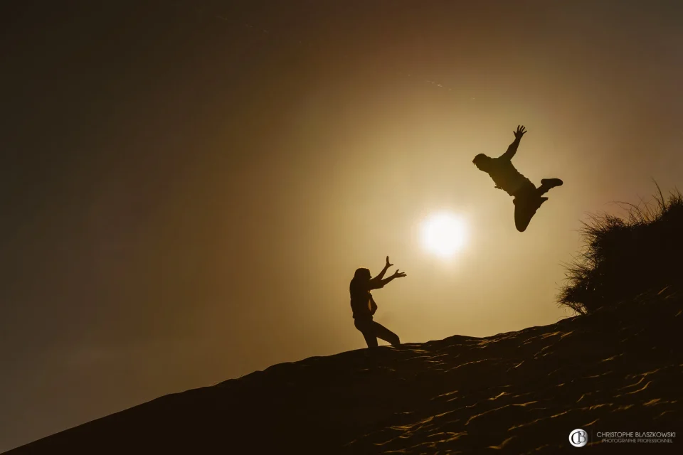 Mariés sur une dune à la plage, la mariée faisant un Kamehameha avec le soleil, créant l’illusion d’une boule de feu, pendant que son mari saute. Photo en contre-jour par Christophe Blaszkowski, photographe de mariage sur Lille.