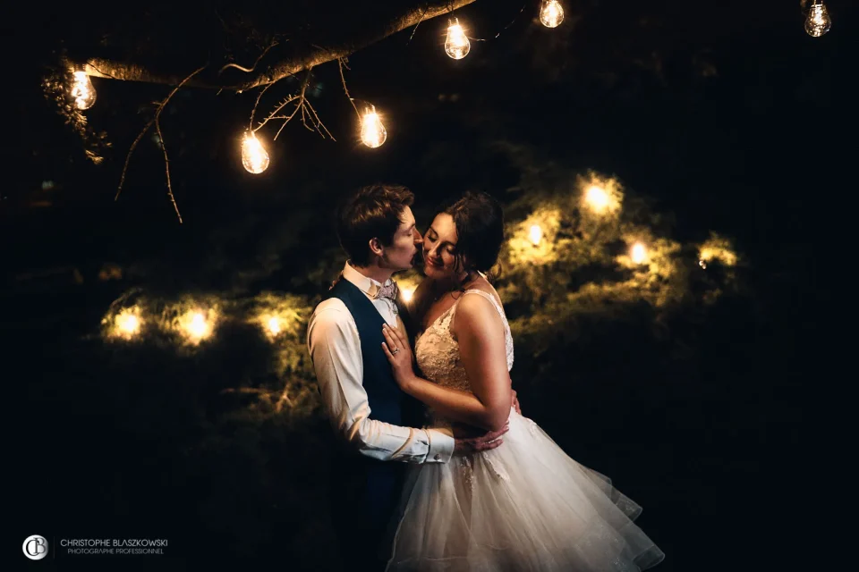 Couple de mariés s’embrassant sous les guirlandes LED accrochées aux arbres, créant une atmosphère féérique et romantique. Photo signée Christophe Blaszkowski, photographe de mariage sur Lille.