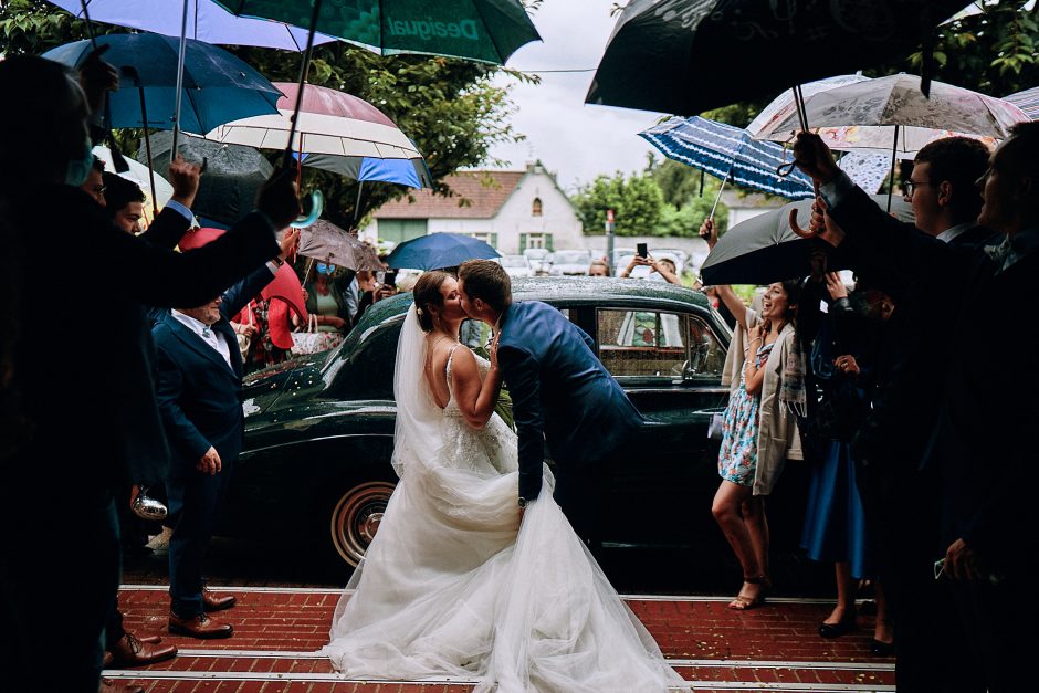 Mariage sous la pluie - les maries s'embrassent et les invites jouent avec les parapluies en sortie de ceremonie