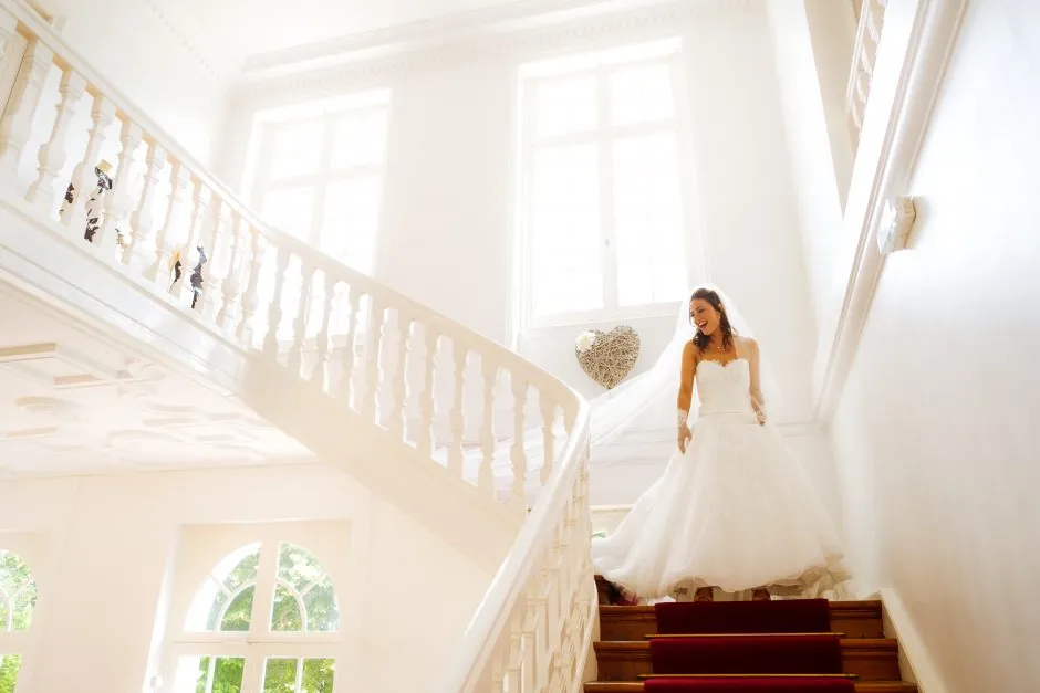 Mariée descendant les escaliers du Château Blanc d'Olivier Sinic, souriant en découvrant son futur mari. La lumière douce et l'intérieur blanc du château ajoutent une touche féérique à ce moment unique. Photo de mariage de Christophe Blaszkowski.