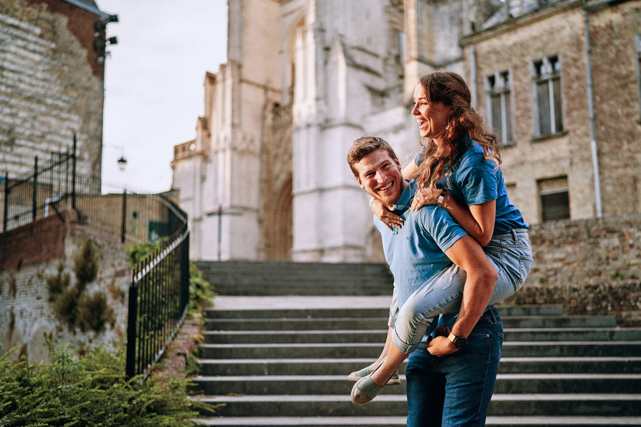 Photo de couple pendant la séance d'engagement avec l'homme portant sa femme sur son dos, riant devant le beffroi de Saint-Omer. Artisan photographe pour votre grand jour. Photographe Mariage Ath - Belgique
