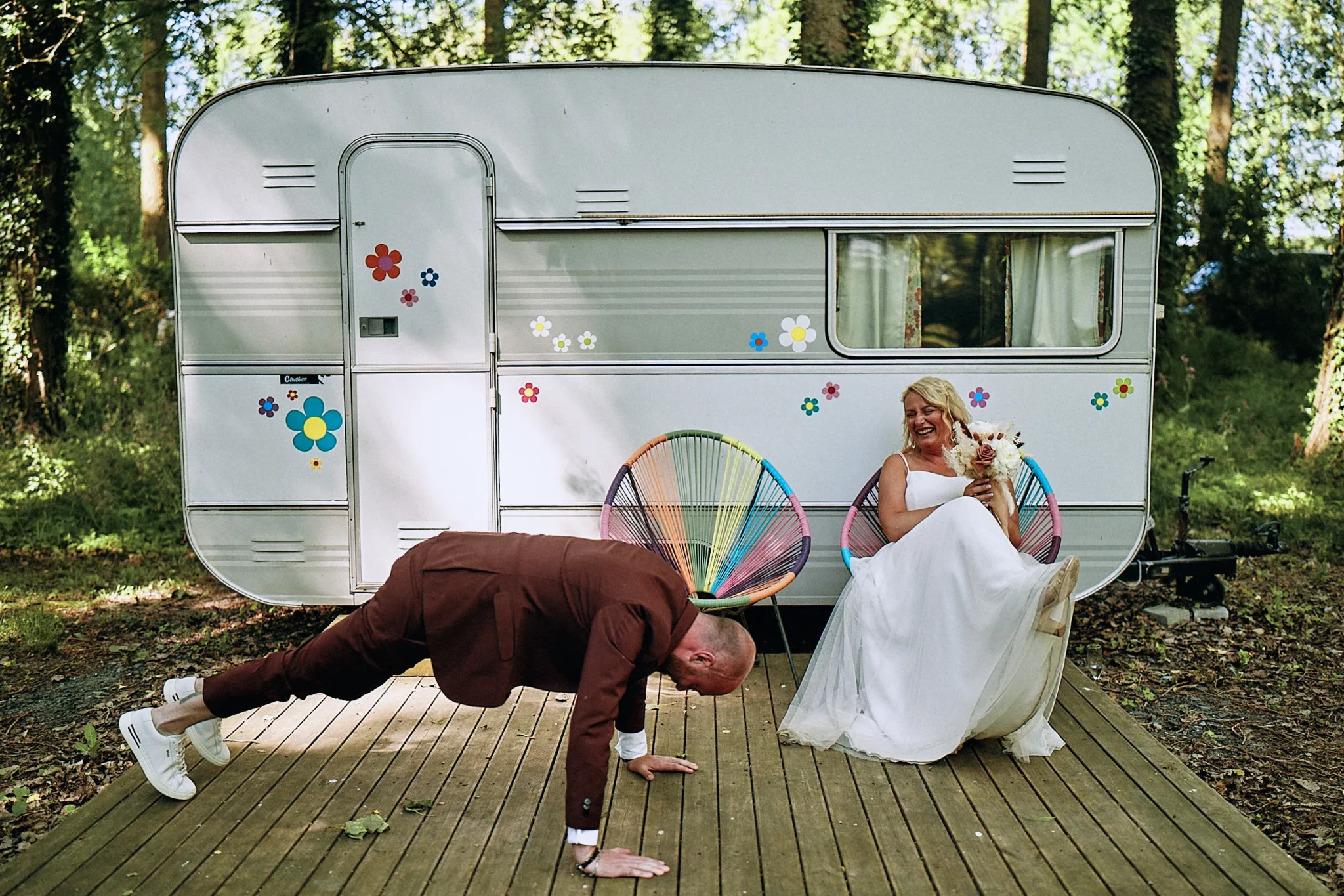 Photo amusante de mariage : le marié fait des pompes pendant que la mariée rit aux éclats, assise sur une chaise. Une caravane en arrière-plan ajoute une touche vintage. Photographe de mariage à Lille : Christophe Blaszkowski. Les mariés sont de Cayeux-sur-Mer