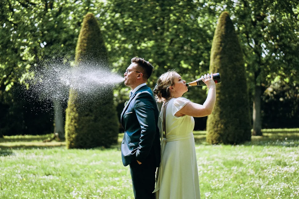 Mariée buvant à la bouteille de champagne et marié recrachant, photo drôle et originale prise de dos au Jardin des Plantes de Lille. Capture signée Christophe Blaszkowski, photographe de mariage sur Lille. Les mariés sont de Hazebrouck