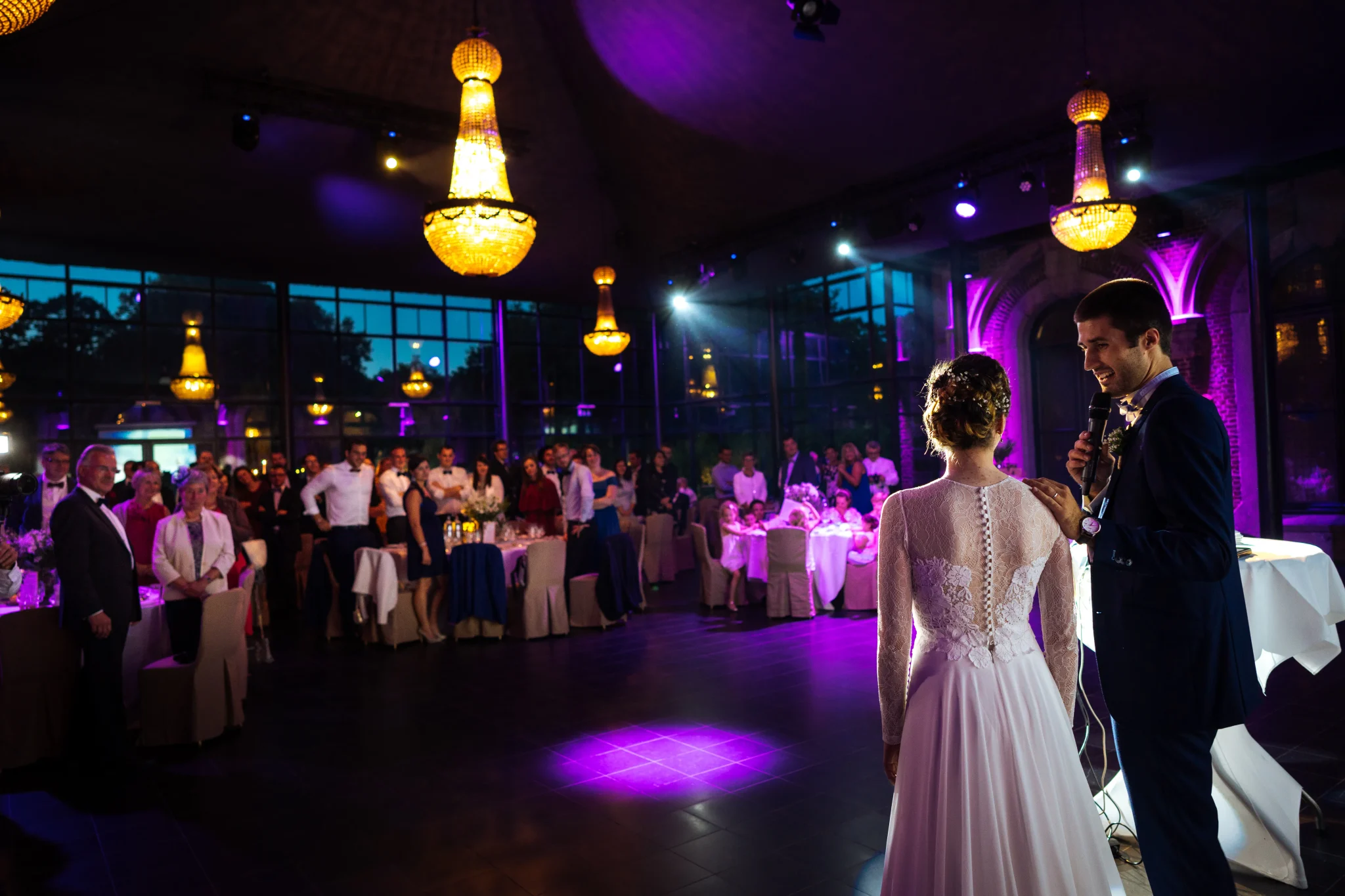 Discours des mariés devant leurs invités sous la belle lumière des lustres du Château de Bourgogne, près de Lille. Le marié pose tendrement la main sur l’épaule de sa femme et la regarde avec amour. Photo capturée par Christophe Blaszkowski, photographe de mariage sur Lille.