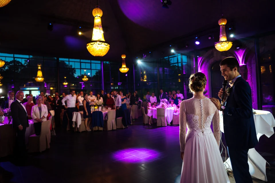 Mariage au Château de Bourgogne à Estaimpuis – Lieu romantique et photogénique 3 Discours des mariés devant leurs invités sous la belle lumière des lustres du Château de Bourgogne, près de Lille. Le marié pose tendrement la main sur l’épaule de sa femme et la regarde avec amour. Photo capturée par Christophe Blaszkowski, photographe de mariage sur Lille.