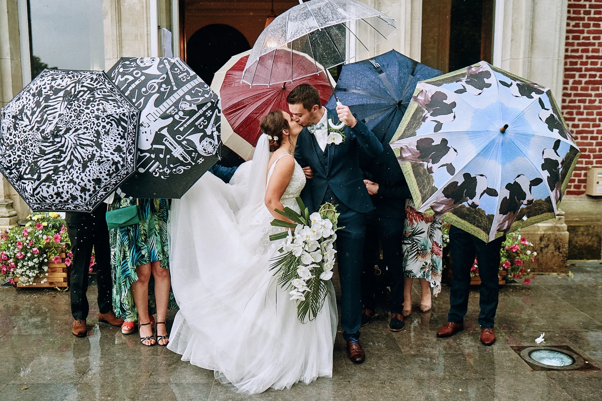 Les mariés de Lys-lez-Lannoy s'embrassant à la sortie de la mairie, entourés d'invités cachés derrière leurs parapluies sous la pluie. Photo de mariage par Christophe Blaszkowski, Spécialiste de la photographie de mariage à Lille