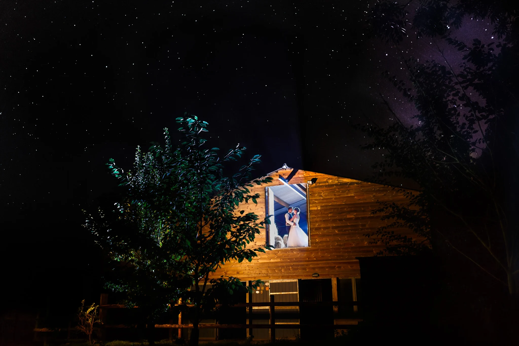 Mariés à l’étage d’une grange à Mons, avec en arrière-plan un ciel étoilé et la structure boisée du lieu. Ambiance romantique et chaleureuse pour une photo de mariage féérique.