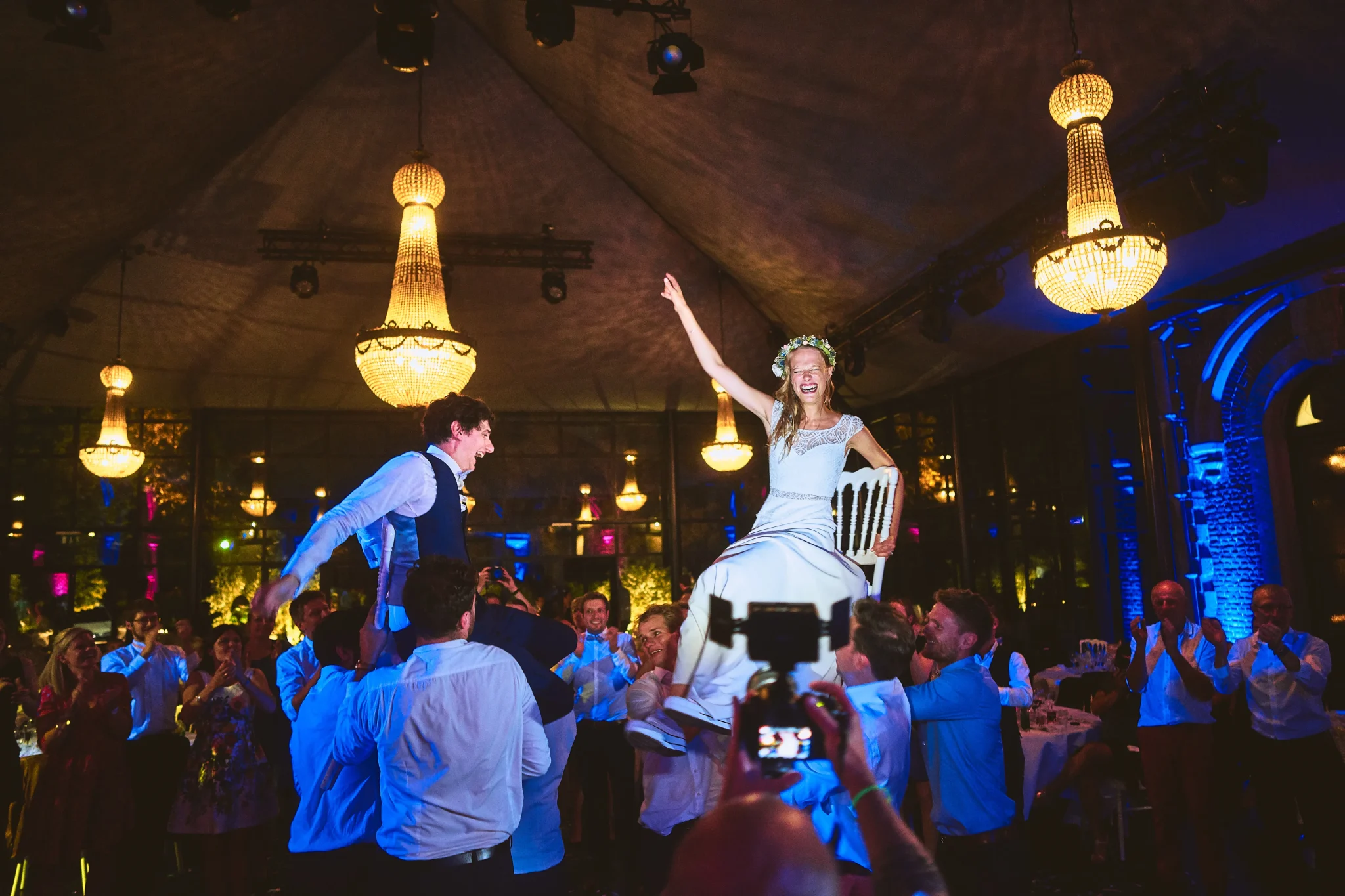 Les mariés de Montataire soulevés par leurs invités pendant la soirée dansante, sous les lumières des lustres du Château de Bourgogne à Estaimbourg. Photo de mariage par L’expert en photographie de mariage Christophe.