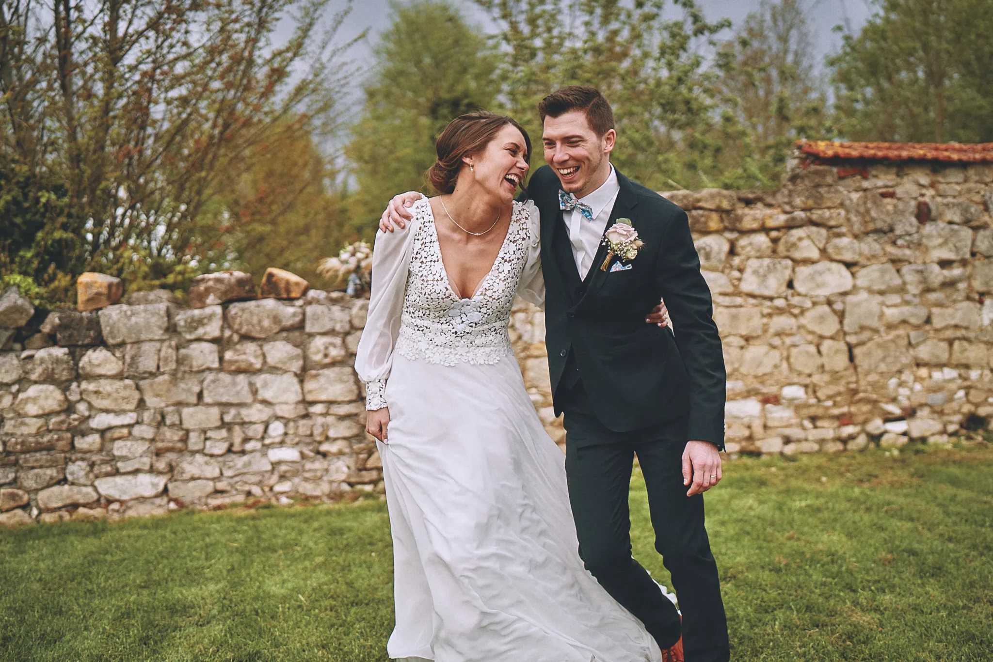 Mariés de Nogent-sur-Oise riant dans le jardin du Domaine La Marlière de Proville, avec un vieux mur de pierre et de la verdure en arrière-plan. Photo de mariage par Christophe Blaszkowski, photographe de mariage sur Lille.