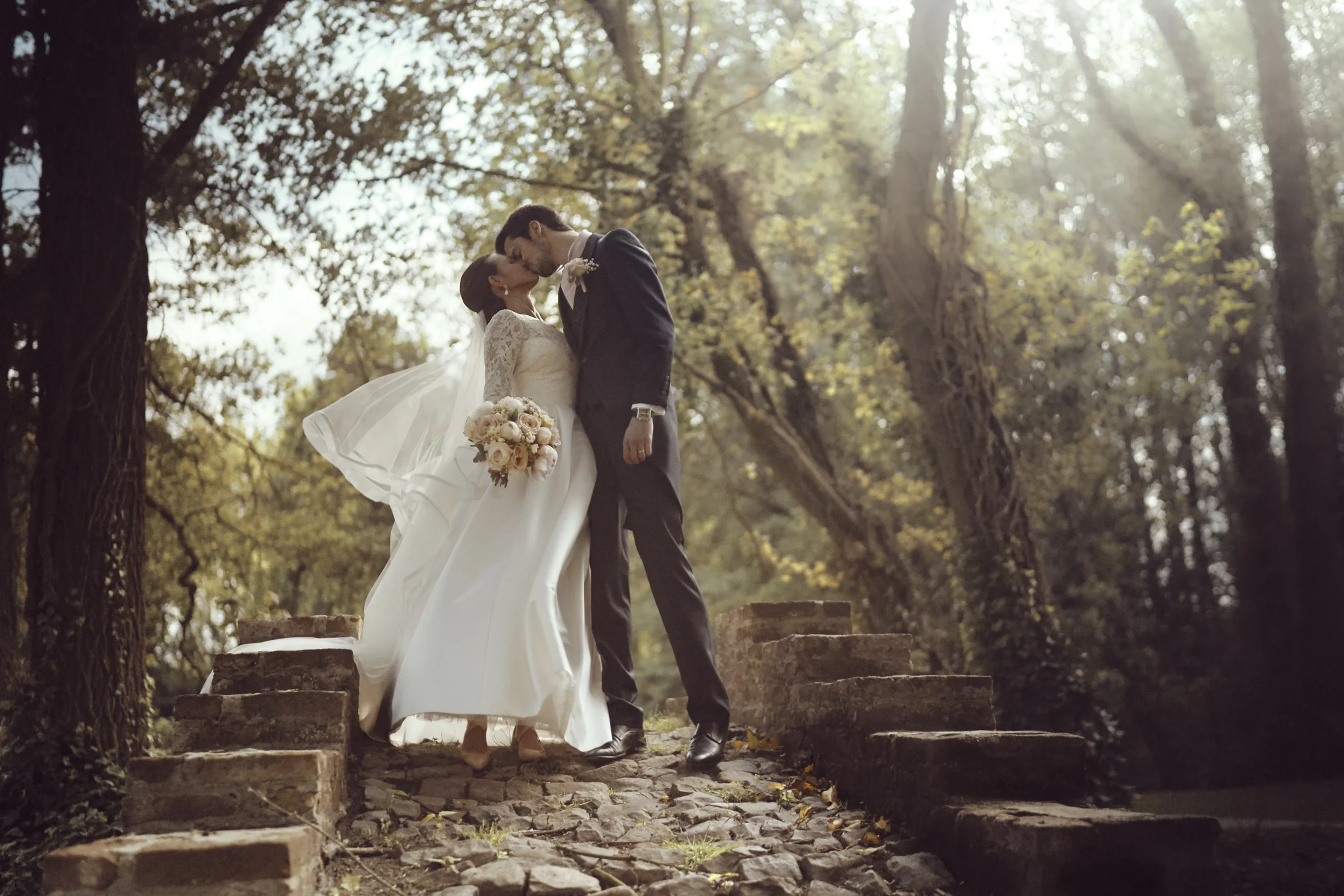 Mariés de Versailles s'embrassant sur le pont du Domaine Clos Barthélémy à Eterpigny, avec les rayons du soleil filtrant à travers les arbres. Photo de mariage par Christophe Blaszkowski, Photographe passionné pour vos souvenirs de mariage sur Lille.