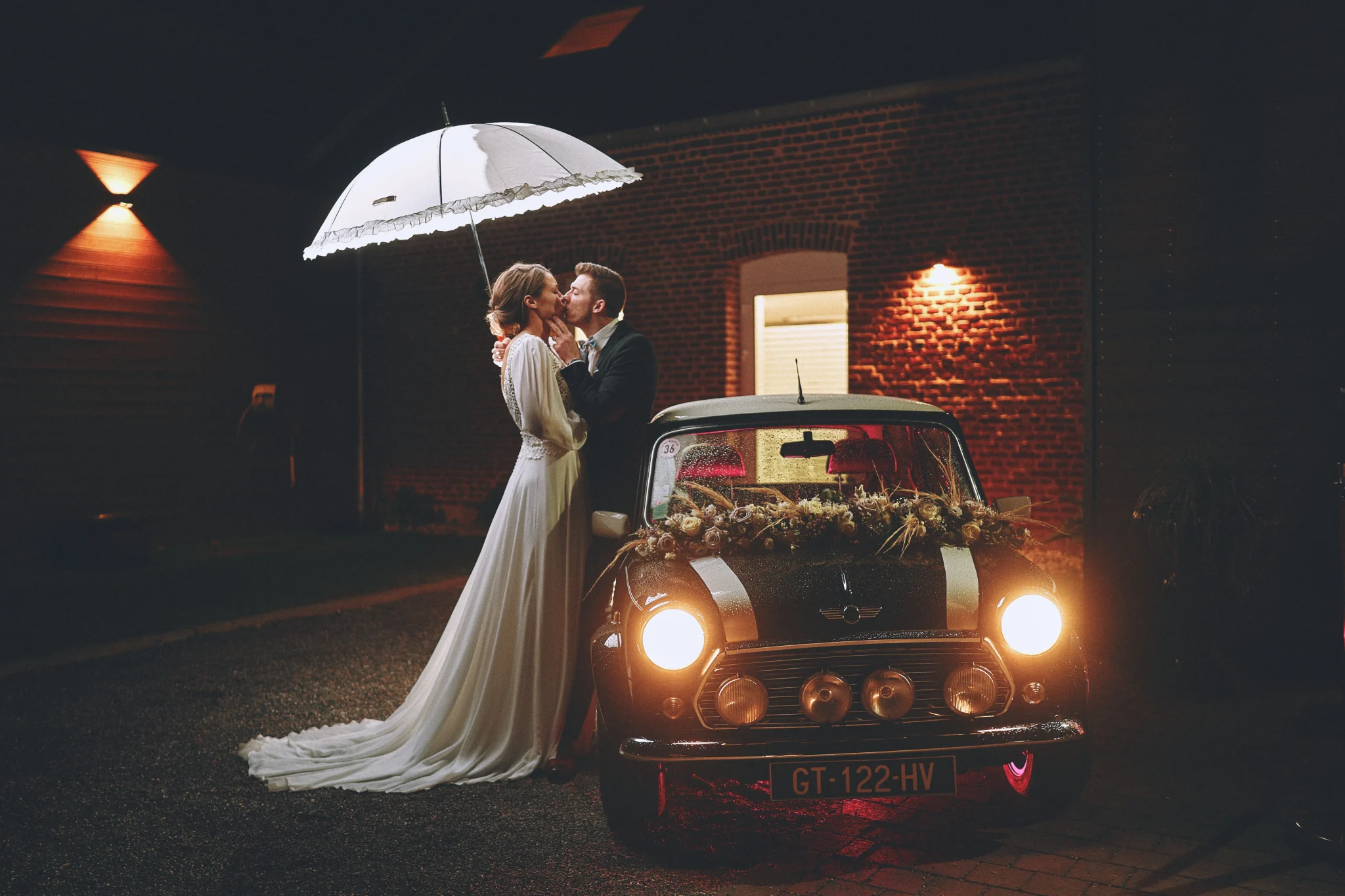 Mariés de l'Oise à côté de leur Mini Austin, photo de nuit avec lumière de la voiture et fond en briques rouges. Photographe de mariage romantique.