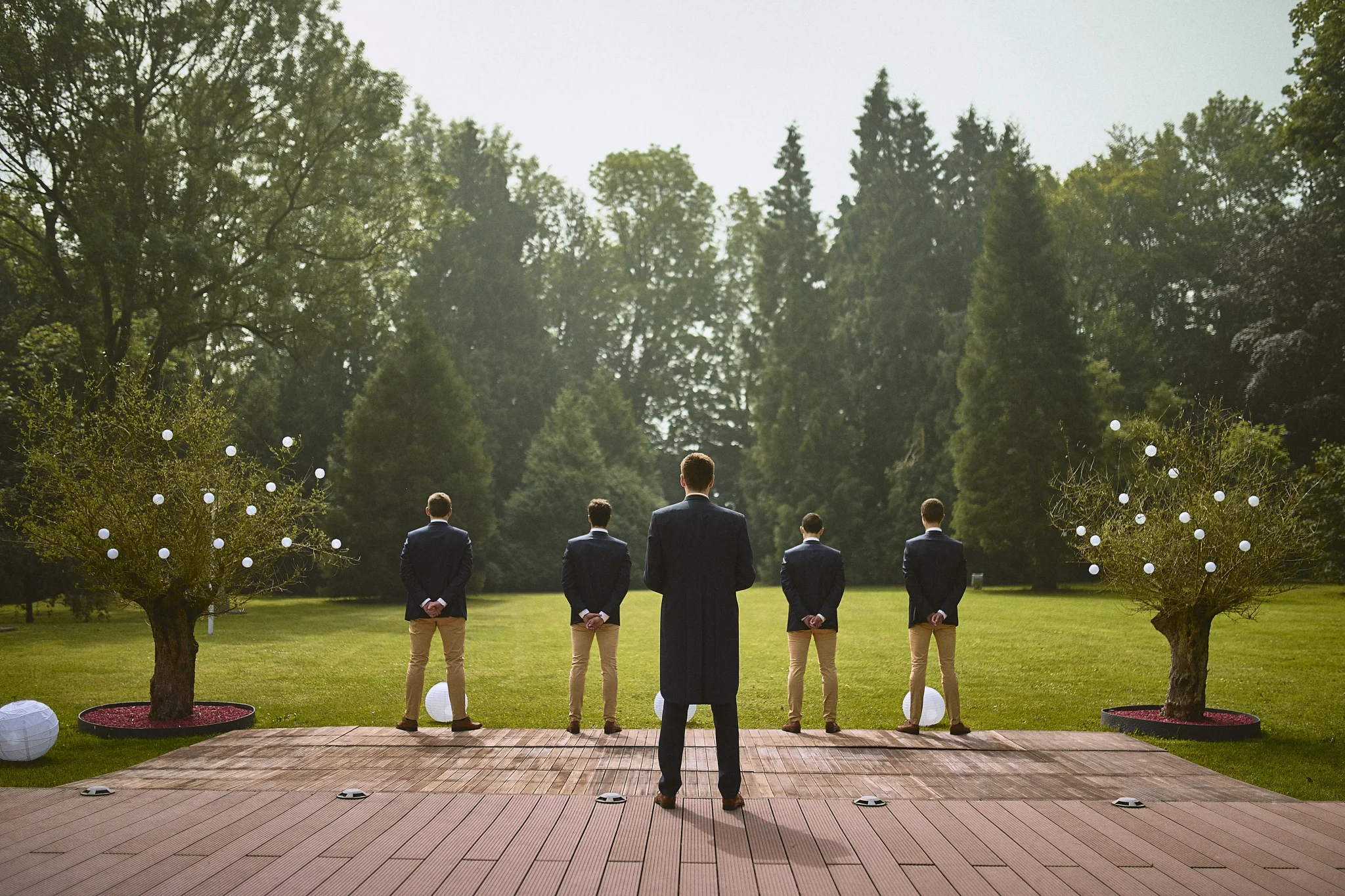 Le marié et ses témoins posant de dos sur la terrasse, attendant l'arrivée de la mariée pour le discours, avec un jardin magnifique en arrière-plan. Photo de mariage par Christophe Blaszkowski, Expert en photographie de couple