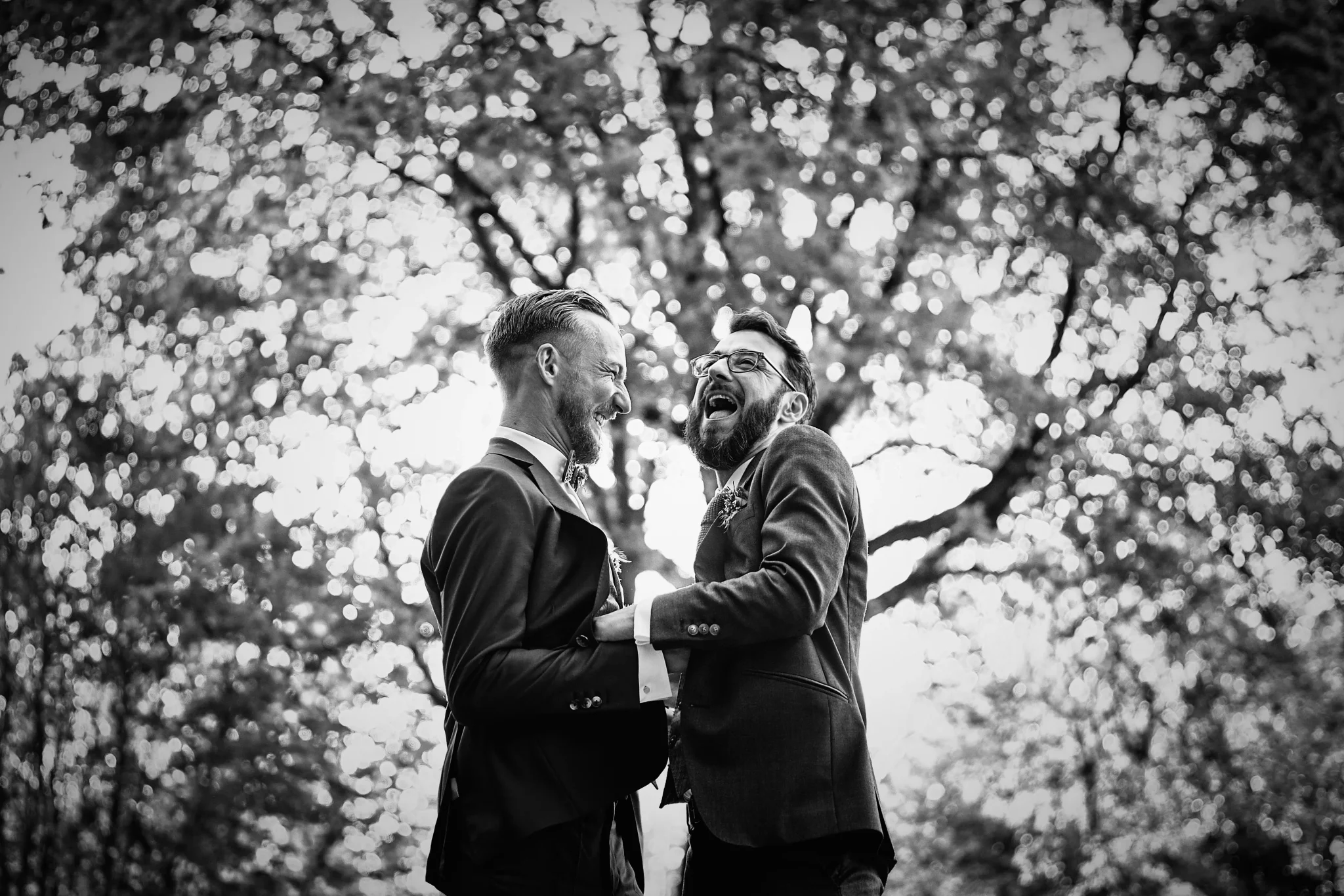 Photo en noir et blanc d’un couple gay souriant, heureux et complice, avec en arrière-plan un arbre LGBTQ+ représentant l’amour et la diversité. Photographe de mariage à Lille : Christophe Blaszkowski.