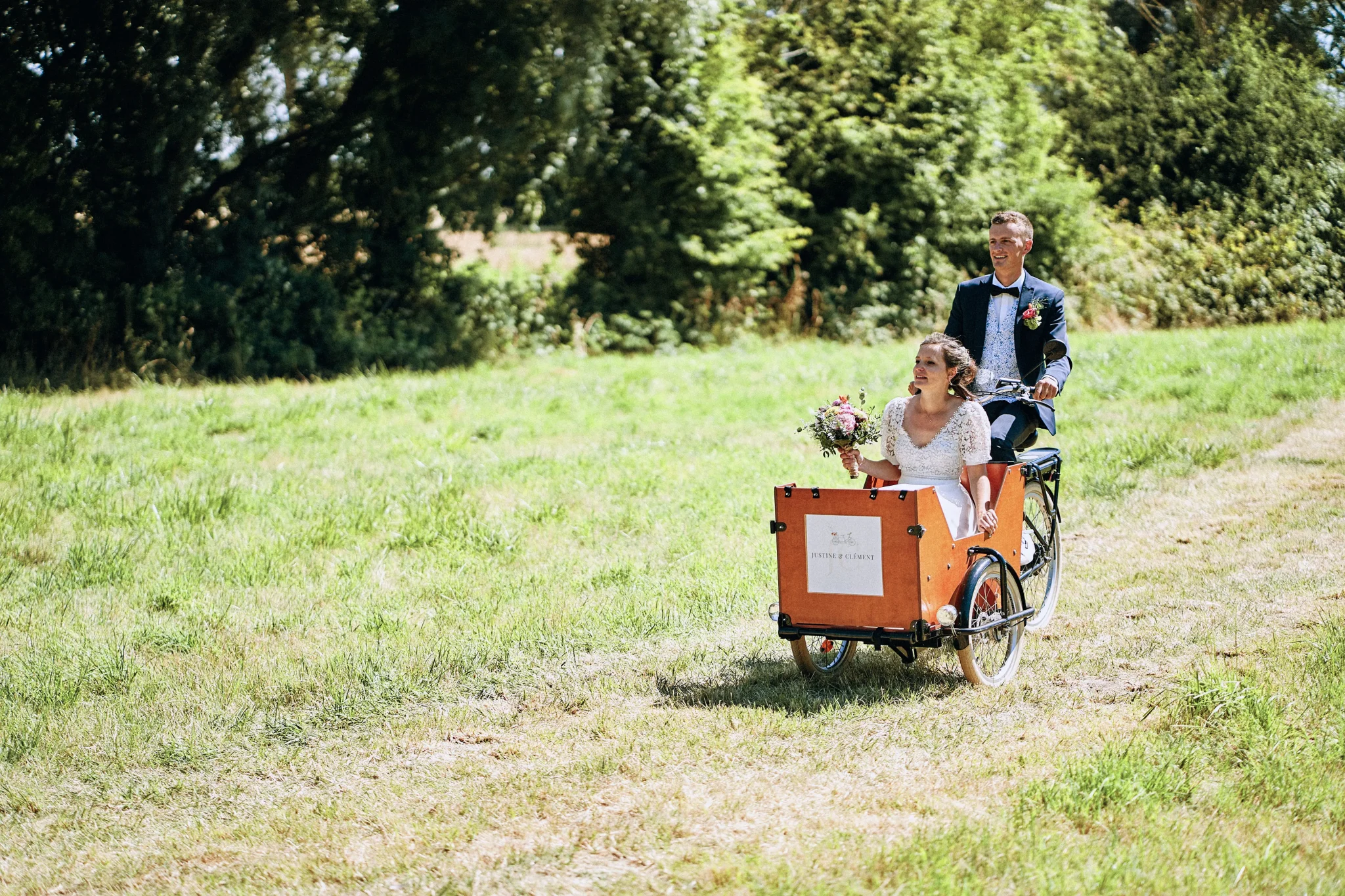 Les mariés de Bruay-la-Buissière arrivant à leur cérémonie laïque dans un tricycle, roulant sur un chemin de prairie. Photo de mariage par Christophe Blaszkowski, photographe de mariage sur Lille.